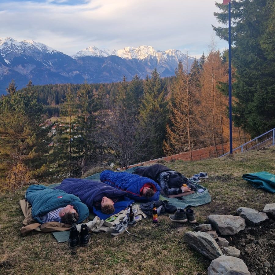 Drei Personen liegen in Schlafsäcken in der Natur mit Bergsicht im Hintergrund.