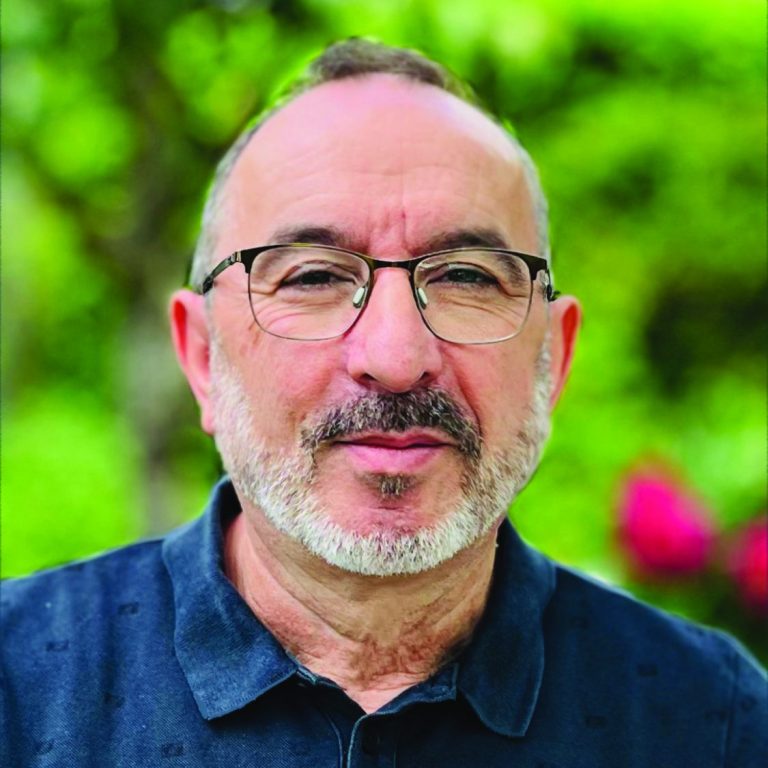 Mann mit Brille und Bart, lächelt vor einer grünen Hintergrundlandschaft.