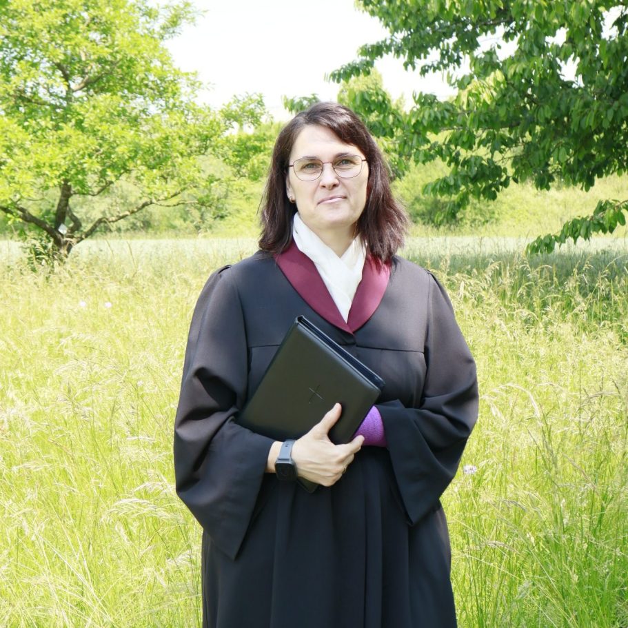 Eine Person in schwarzem Talar mit einem Ordner, stehend in einer grünen Wiese.
