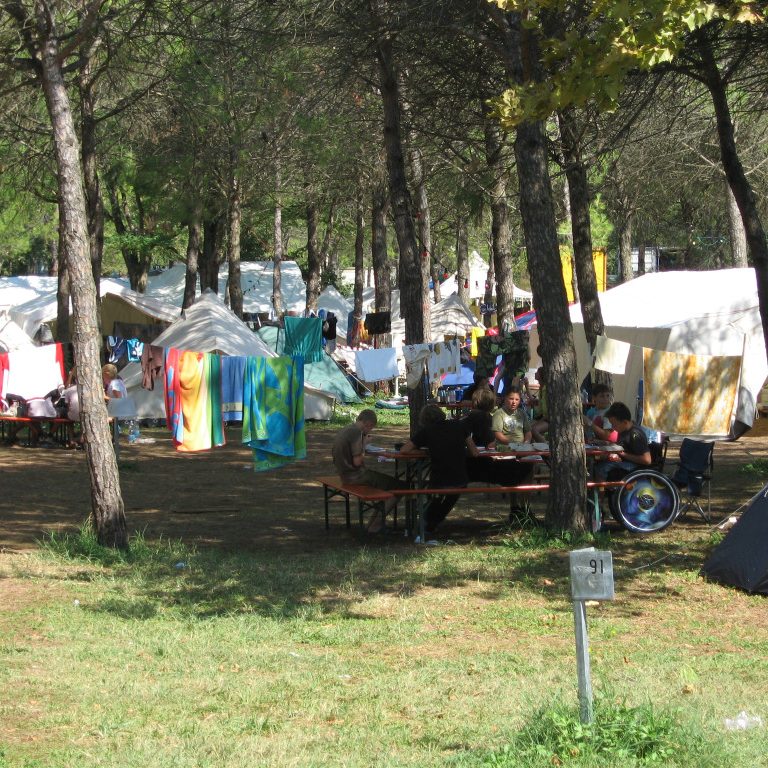 Zeltlager mit vielen Zelten und Biertischgarnituren unter Bäumen in einer grünen Wiese.