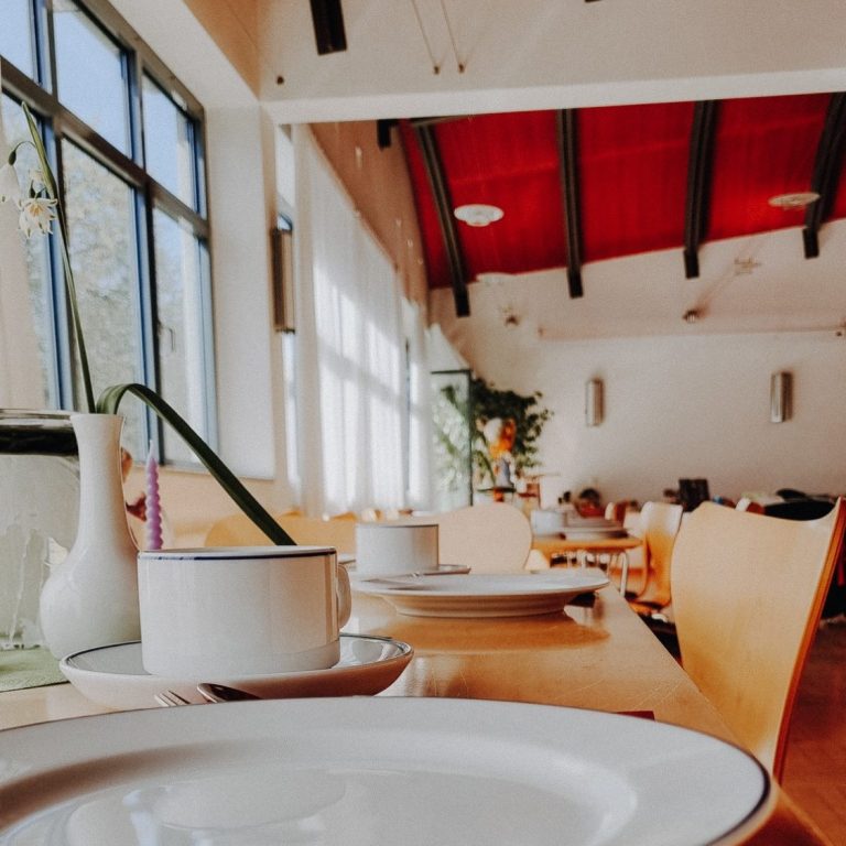 Helles Interior mit Tischen, Geschirr und Pflanzen im Hintergrund.