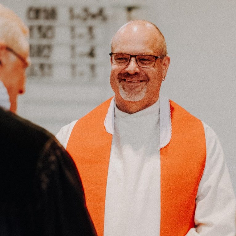 Ein lächelnder Mann in weißem Talar mit orangefarbener Stola steht vor einer Person.