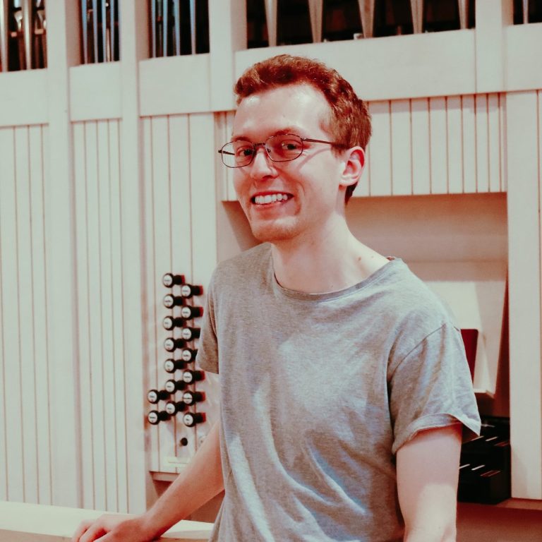 Junger Mann mit Brille steht vor einer Orgel in einem hellen Raum.