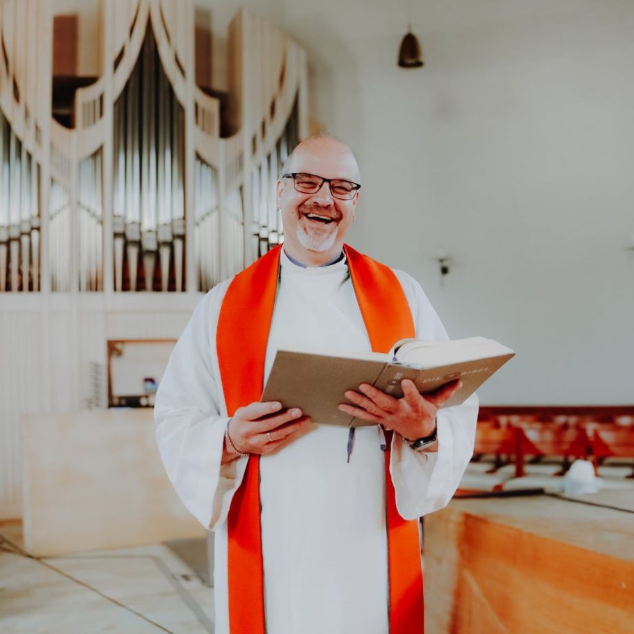 Ein lächelnder Diakon hält eine Bibel und trägt eine orangefarbene Stola und einen weißen Talar in einer Kirche.