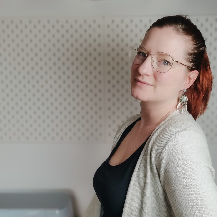 Frau mit Brille und roten Haaren, lächelt leicht vor einer neutralen Wand.