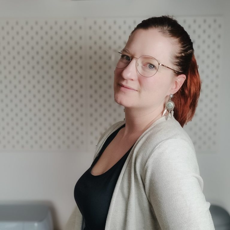 Frau mit roten Haaren und Brille, lächelnd, in einem hellen Raum.
