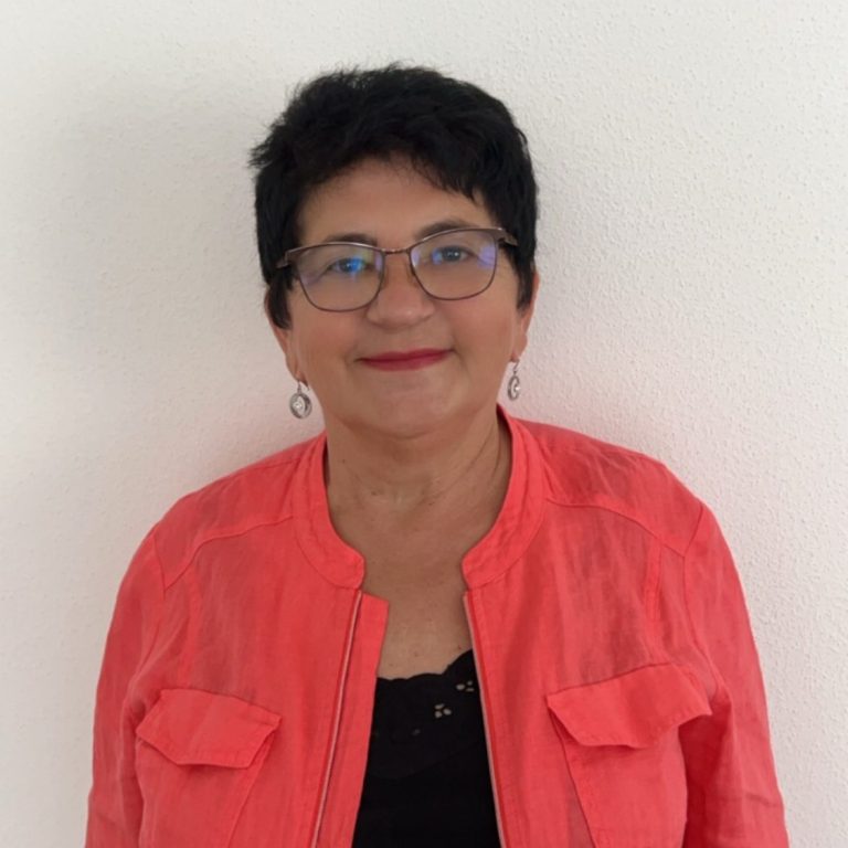Ältere Frau mit schwarzem Haar, Brille und einem korallfarbenen Blazer, lächelt freundlich.