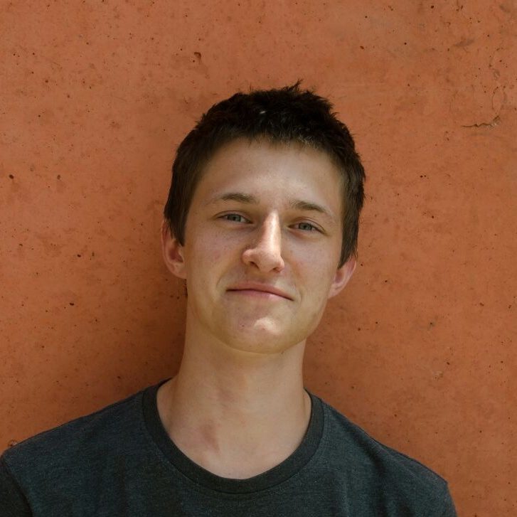 Junger Mann mit kurzer Frisur vor orangefarbener Wand, lächelt freundlich.