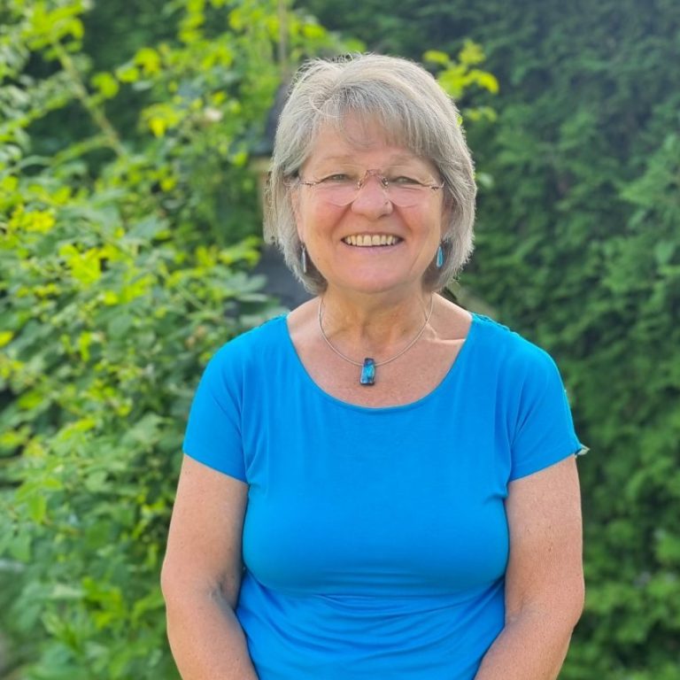 Ältere Frau mit grauen Haaren, trägt ein blaues Shirt, lächelt vor grüner Kulisse.