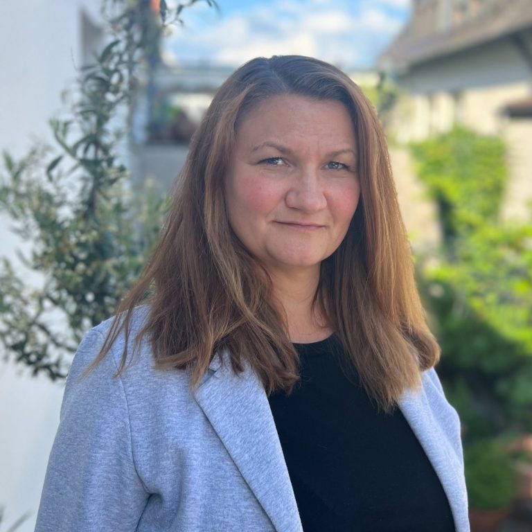 Frau mit langen braunen Haaren, trägt eine graue Jacke und steht im Freien.