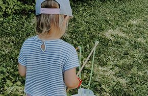 Ein kleines Kind mit einer Kappe hält Spielzeugstäbe und steht auf Gras.